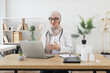 © sofiko14 - Young Muslim female doctor having online consultation using laptop. Asian woman in professional medical setting with stethoscope discussing with patient. Concept of telemedicine and healthcare.