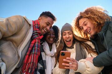 four young cheerful multi ethnic people having fun together outdoors in winter, looking at smartphon