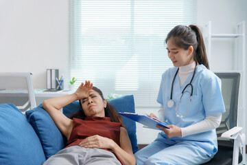 Wall Mural - Young female doctor visiting sick mature woman lying on sofa and holding medical chart writing prescription