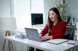 © Apichat - A woman is sitting at a desk with a laptop and a stack of papers