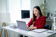 © Apichat - A woman is sitting at a desk with a laptop and a stack of papers