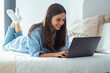 © simona - Smiling young woman typing on laptop while lying on bed, happy girl using computer for chatting writing post in social network, browsing web, buying online or internet blogging at home