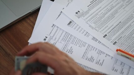 Poster - Businessman holds in hands fan of dollar banknotes in front of W-4 1040 W-9 tax forms, person sets workplace with laptop and stack of documents slow motion