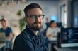 © Mediaphotos - Portrait of adult bearded man as IT developer wearing glasses sitting by desk in modern office and looking at camera with people working in blurred background copy space