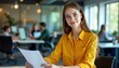 © Maryna - Young woman sits at desk in modern office. Wears yellow shirt, looks at camera. Holds papers in hands. People work in background. Busy office environment with computers, documents. Woman appears