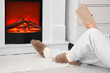 © Pixel-Shot - Woman in soft slippers reading book near fireplace at home