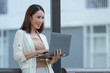 © crizzystudio - Young asian businesswoman is standing outdoors in an urban setting, smiling as she uses her laptop, dressed in business casual attire, suggesting a modern and flexible work style