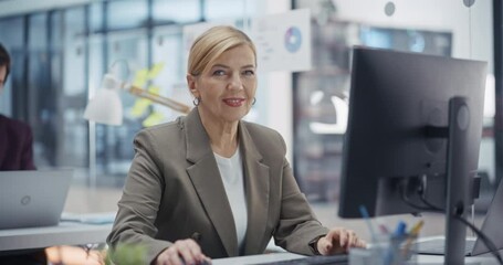Aufkleber - Middle Aged Successful Caucasian Woman Looking at Camera and Smiling. Professional Female Project Manager Working on Desktop Computer in Office. Colleague Working in the Background