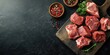 © Lala - Fresh uncooked red meat cuts arranged on a dark wooden cutting board with peppercorns in wooden bowls and fresh herbs on a textured black surface.