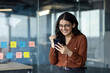 © Liubomir - Successful woman at workplace standing with phone in hands. Businesswoman celebrating victory and successful achievement results. Office worker received online notification message on smartphone.