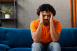 © ViDi Studio - Young sad sick ill Indian man wear orange casual clothes sits on blue sofa couch put hand on head suffer stay at home hotel flat rest relax spend free spare time in living room indoor. Lounge concept.