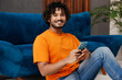 © ViDi Studio - Young smiling Indian man wear orange casual clothes sit near blue sofa couch hold use mobile cell phone stay at home hotel flat rest relax spend free spare time in living room indoor. Lounge concept.