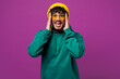 © ViDi Studio - Young happy shocked surprised excited Indian man he wear green hoody yellow hat sunglasses casual clothes hold head look camera isolated on plain purple background studio portrait. Lifestyle concept.