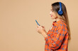 © ViDi Studio - Side profile view young woman wear orange shirt casual clothes listen to music in headphones use mobile cell phone isolated on plain pastel light beige background studio portrait. Lifestyle concept.