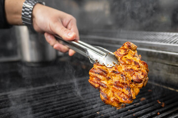 Wall Mural - A close-up of a hand holding tongs, flipping a large, juicy piece of grilled meat on a smoky barbecue grill. The image captures the essence of outdoor cooking and delicious food preparation.