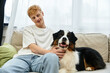 © LIGHTFIELD STUDIOS - A young redhead man relaxes on a couch, lovingly petting his Australian Shepherd dog at home.
