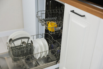 Side view of an open dishwasher with dishes and cutlery against a background of white wooden cabinets. Dishwashing concept, comfort. High quality photo