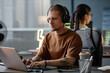 © Seventyfour - Medium shot of confident male IT developer working in headphones immersed in coding on laptop at cluttered desk in corporate office, copy space