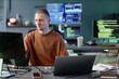 © Seventyfour - Medium shot of calm male programmer typing on computer keyboard working alone at cluttered desk with laptop, captured against screens displaying lines of code in tech company office