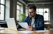 © NS Frames - Man sitting at a wooden table in a modern office environment. He appears to be deep in thought as he examines a document in his hand while looking at a laptop in front of him