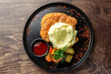 Wall Mural - A delicious serving of crispy fried chicken with creamy mashed potatoes, accompanied by a side of mixed vegetables, pickles, and a small bowl of ketchup, all on a black plate.