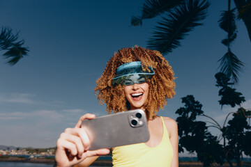 Sticker - Young woman smiling and taking a selfie by the beach, wearing a stylish visor hat in bright sunlight