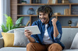 © Liubomir - Man sitting with tablet computer at home on sofa, wearing headphones, watching online sports match, celebrating successful results achieved in video game stream.