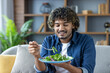 © Liubomir - Joyful and satisfied man eating salad, diet food while sitting on sofa in living room at home. Proper nutrition and vegetarian food.