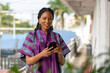© Roger - Smiling woman in vibrant African print dress uses her phone outdoors.