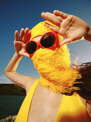 Poster - Woman, sunglasses, yellow hat, beard, water, blue sky a woman in sunglasses and a yellow hat with a beard on top of her head stands by the water, framed by a clear blue sky