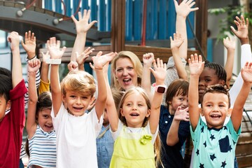Poster - Group of diverse children, boys and girls, with Caucasian woman teacher, smiling and raising hands. Happy children and teacher in a playground. Diverse student with teacher raising hand in playground