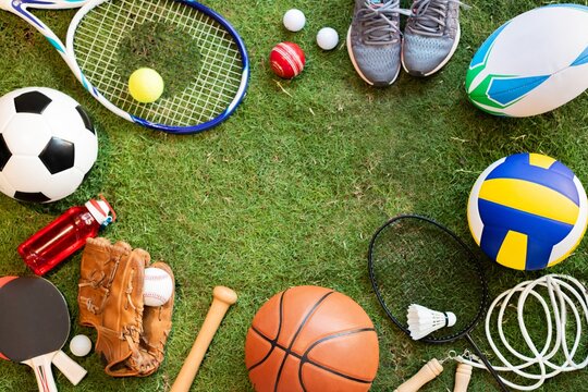 Various sports equipment on grass, including a soccer ball, tennis racket, and basketball. Sports gear like shoes and gloves complete the athletic theme. Sport background, with grass field background.