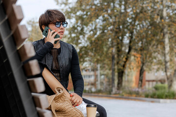 Brunette with short hair, sitting on a bench in a city park, holding a phone, talking, gesticulating, drinking coffee from a cardboard cup, on a summer day.