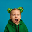© master1305 - Portrait of emotive child, girl in green hoodie and buns hairstyle shouting, expressing anger and dissatisfaction against blue studio background. Concept of childhood, emotions, lifestyle