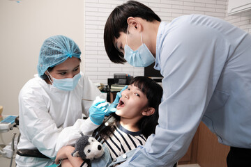 Wall Mural - Asian female pediatric dentist examines a girl's teeth in dental clinic and takes care and encourages of father, mouth oral hygiene, and professional orthodontic healthcare work in a kid hospital.