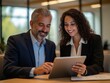 © Arip - Team of diverse partners mature Latin business man and European business woman discussing project on tablet sitting at table in office.