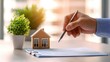© weerasak - A hand holds a pen over a document next to a small house model and a potted plant, suggesting real estate or home planning activities.