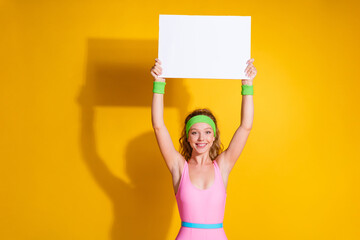 Wall Mural - Young woman in retro sportswear holding blank placard on yellow background, smiling energetically, representing fitness and vintage fashion