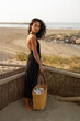 © sutulastock - Brunette Woman in Black Dress Standing and Looking at the Beach with Straw Bag