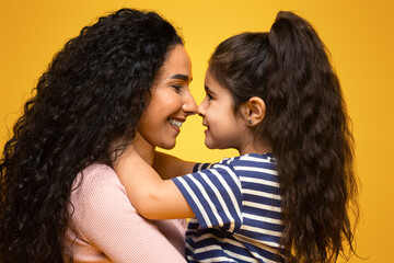 Wall Mural - Love My Mommy. Cute Little Girl Bonding With Her Young Mom, Happy Middle Eastern Mother And Daughter Embracing And Touching Noses While Posing Together Over Yellow Studio Background, Copy Space