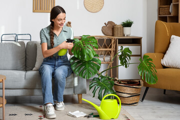 Wall Mural - Young woman spraying water on Monstera houseplant in living room