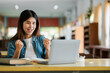 © Satori Studio - A joyful young woman celebrates success while working on a laptop in a bright library, surrounded by books and natural light.