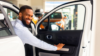 Wall Mural - Happy Black Guy Posing In His New Car And Smiling At Camera, Joyful Young African American Man Opening Door Of Luxury Vehicle In Dealership Center, Purchasing New Automobile, Closeup Shot