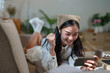 © crizzystudio - Happy young woman is lying on the sofa at home, raising her fist in celebration while playing mobile games on her smartphone, experiencing a moment of joy and excitement
