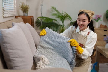 Wall Mural - Young smiling housekeeper wearing yellow gloves is arranging a blanket on a beige sofa in a living room, performing her cleaning duties with joy and dedication