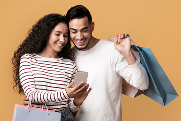 Poster - Discount App. Cheerful Arab Couple Holding Smartphone And Colorful Paper Shopping Bags, Middle Eastern Spouses Browsing Mobile Application With Coupons, Promo Codes, and Deals Over Yellow Background