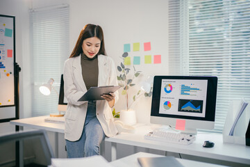 Aufkleber - Young asian businesswoman reviewing financial reports and analyzing statistics on clipboard and computer screen, working in bright modern office