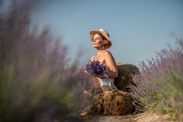 Wall Mural - woman sitting in a field of lavender and wearing a straw hat. She is smiling and holding a bouquet of flowers. Scene is peaceful and serene, as the woman is surrounded by the beauty of nature