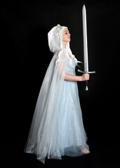 Wall Mural - full length portrait beautiful blonde fantasy maiden faery wearing medieval fairytale ballgown costume, flowing white hooded cape. standing pose holding a sword weapon, isolated dark studio background