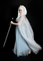 Wall Mural - full length portrait beautiful blonde fantasy maiden faery wearing medieval fairytale ballgown costume, flowing white hooded cape. standing pose holding a sword weapon, isolated dark studio background